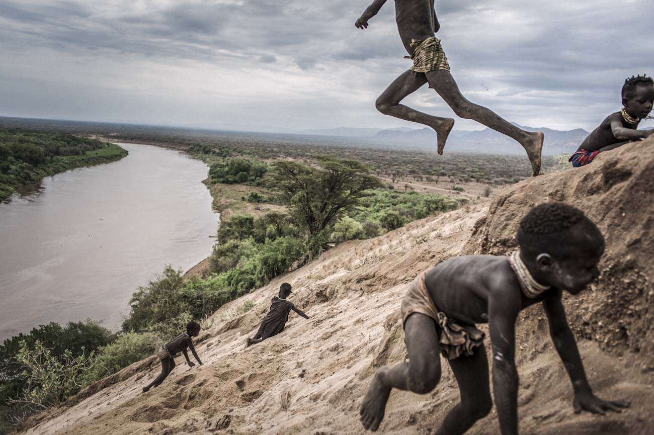 South Ethiopia. 2011. Omo Valley. Along the oriental shore of the Omo River near the Karo village, children are playing by jumping in the sand. The Karo village is located in a natural bights of the Omo River. Karos are a small tribe with an estimated population between 1,000 and 3,000 people and lives thanks to fishing and cultivation made possible by the flooding of the Omo River. Currently, the river forest seen in the background has been demolished and replaced by an extensive cotton plantation of a foreign private company. Following the construction and commissioning of the Gibe III dam, the flooding of the Omo River has stopped, depriving the Karo population of the possibility of cultivating those products that today they’re forced to buy at the market.