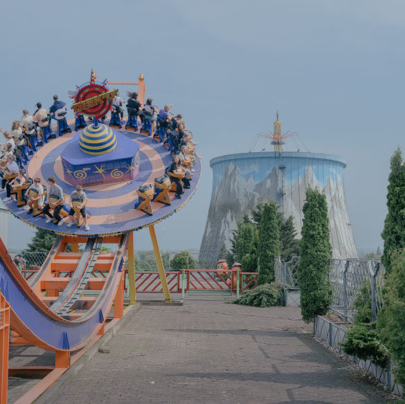 Photographie en plan moyen d'un manège de parc d'attractions, avec une structure en forme de disque bleu et jaune pivotant sur des rails orange, remplie de personnes assises. En arrière-plan, une grande tour de refroidissement cylindrique, décorée d'une fresque montagneuse, se dresse sous un ciel clair.