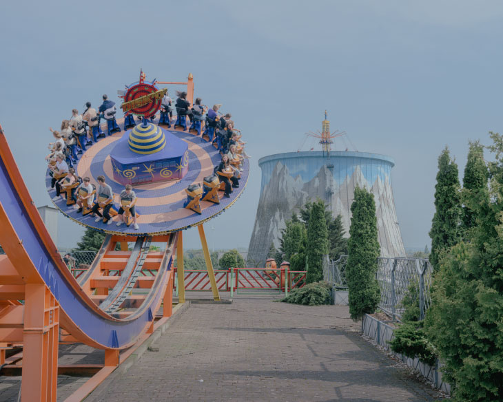 Photographie en plan moyen d'un manège de parc d'attractions, avec une structure en forme de disque bleu et jaune pivotant sur des rails orange, remplie de personnes assises. En arrière-plan, une grande tour de refroidissement cylindrique, décorée d'une fresque montagneuse, se dresse sous un ciel clair.