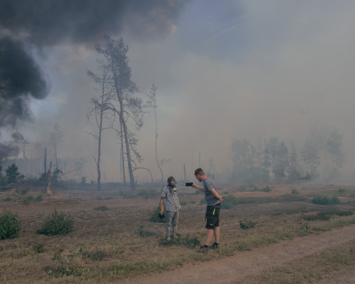 Photographie en plan moyen de deux personnes se tenant dans un champ dévasté et brûlé, avec des troncs d'arbres calcinés et une végétation éparse. Une personne porte un masque à gaz et un t-shirt à carreaux, tandis que l'autre tient un smartphone pour prendre une photo ou une vidéo. De la fumée noire s'élève d'un feu au loin sous un ciel couvert.